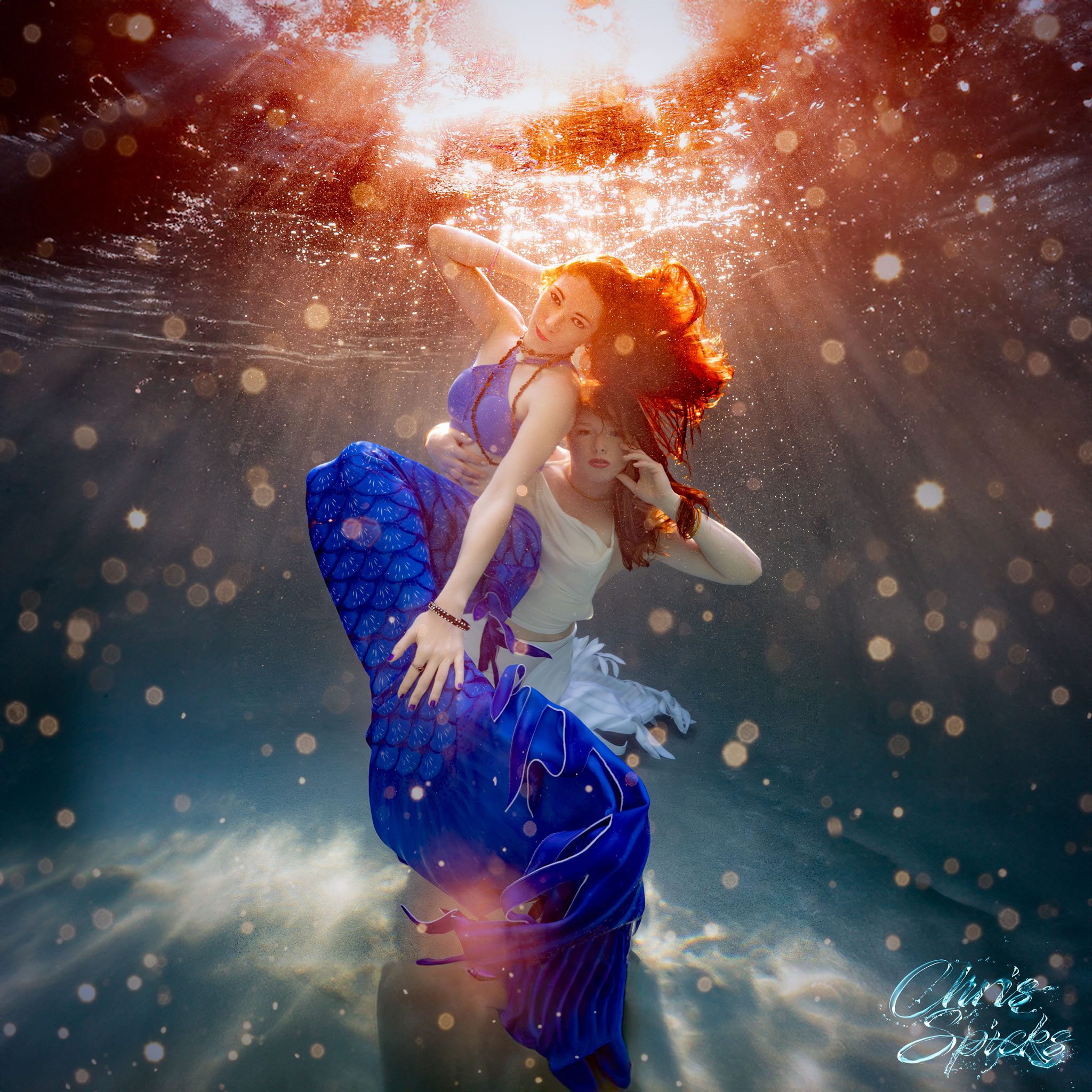 Two women, one dressed as a mermaid in a blue tail and the other in white, swim underwater with sunlight filtering through, creating a magical atmosphere.