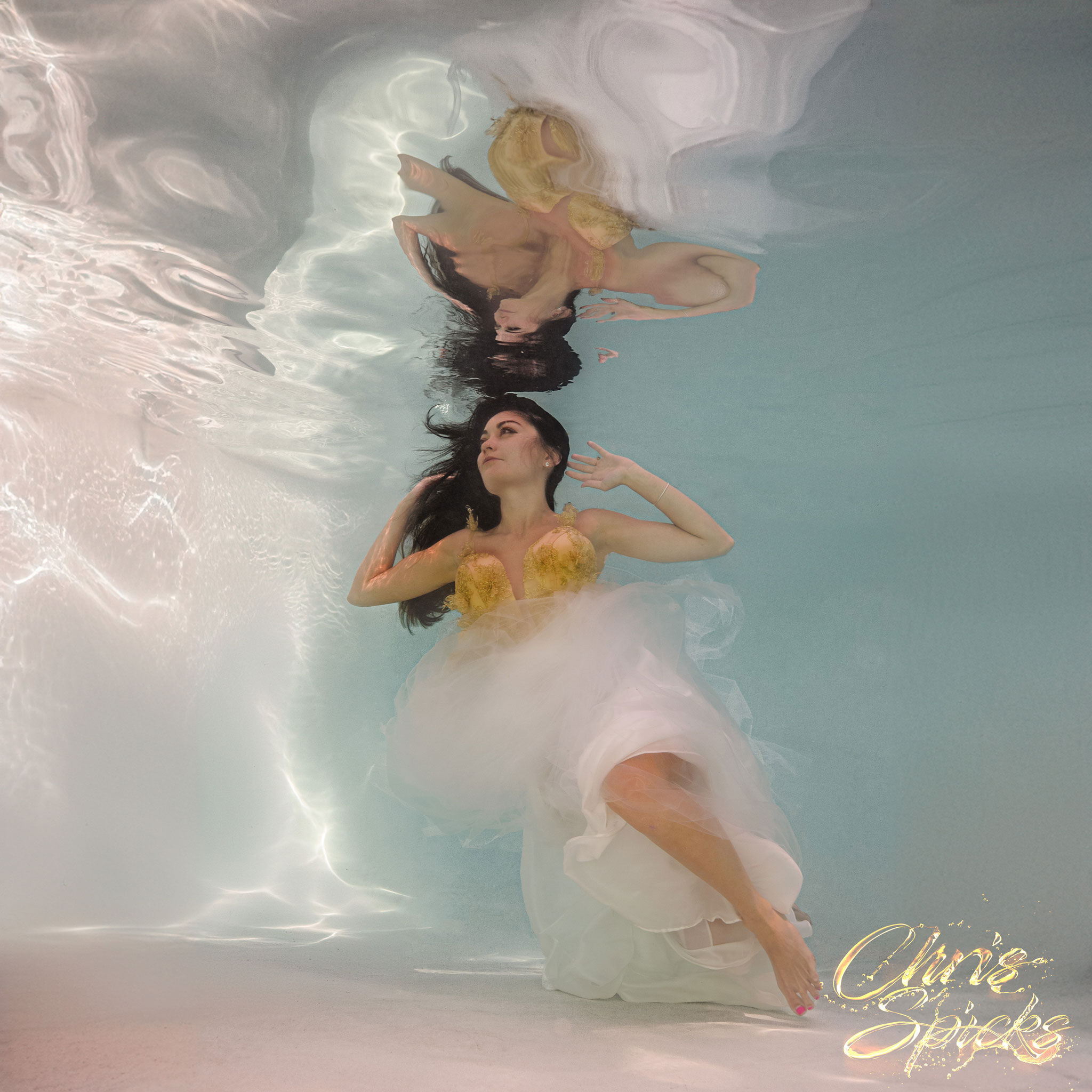 A woman in a yellow and white dress floats underwater, her reflection visible on the water's surface.