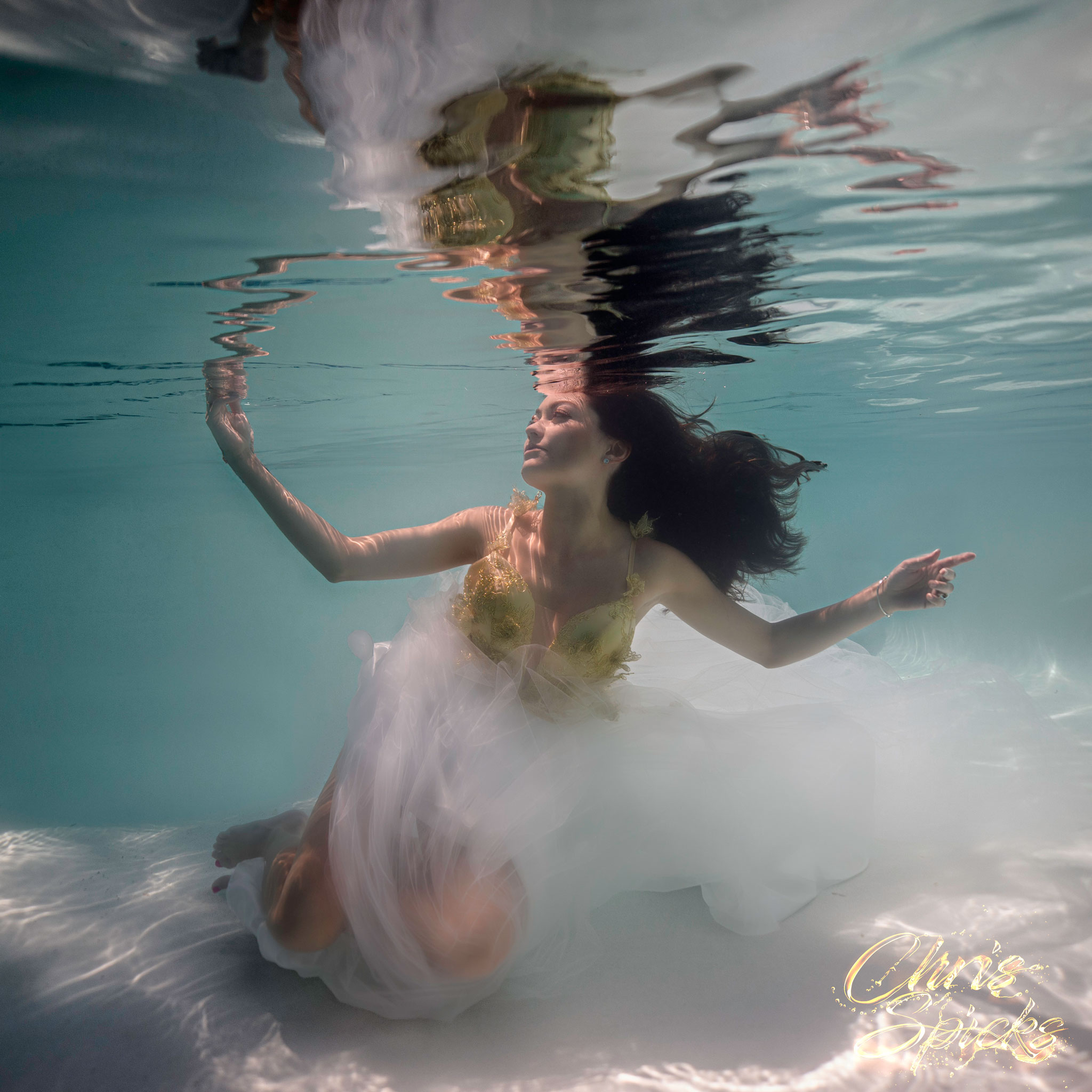 A woman in a flowing white dress and gold bodice is submerged underwater, her hair floating around her as she reaches upward.