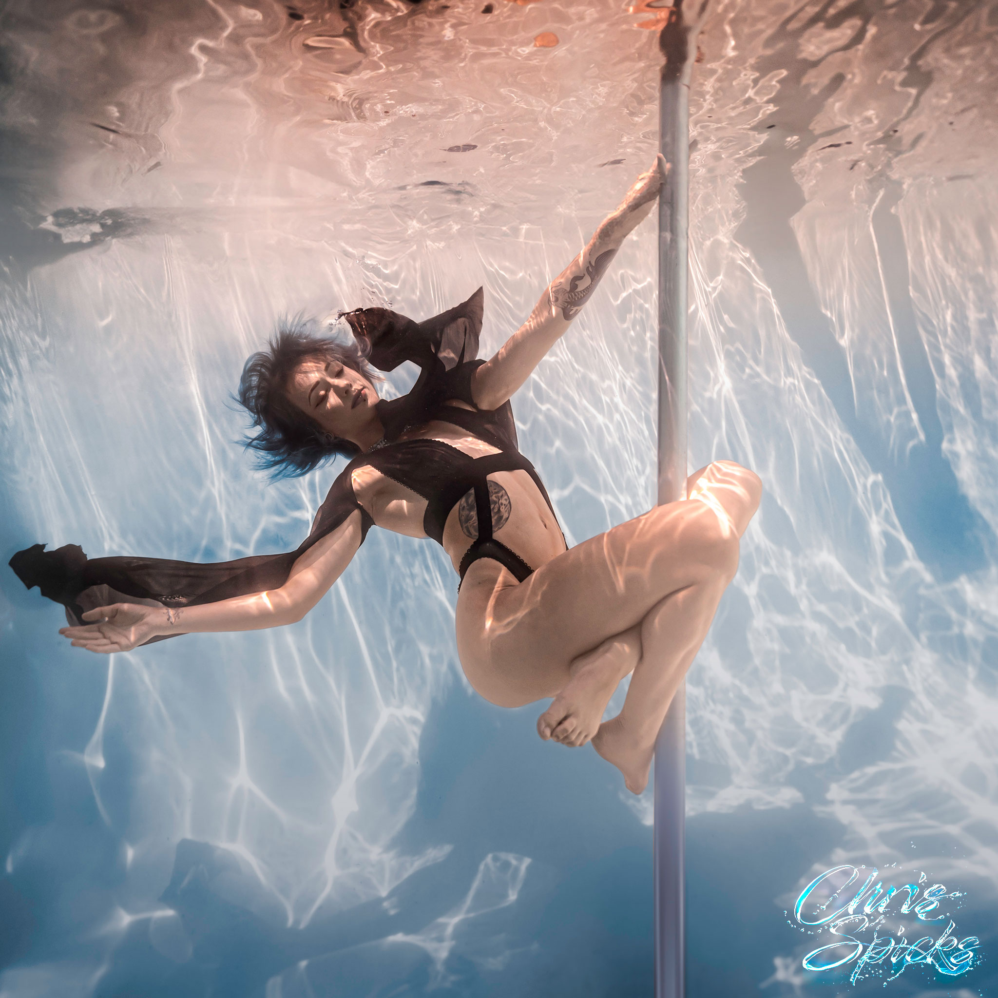 A person performs an underwater pole dance in a pool, wearing a black outfit with flowing sleeves.