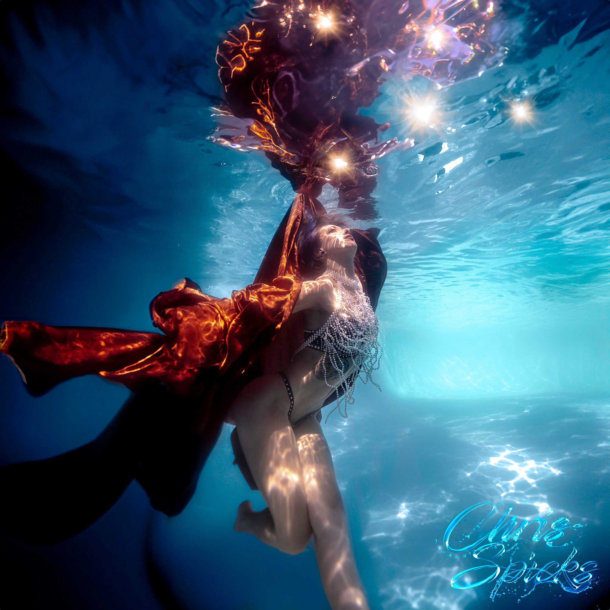 A person underwater in a revealing outfit and ornate headgear, reaching upward with a glowing object. The water reflects pale yellow lights below and blue hues around. "Omer Sipahi" text in the corner.