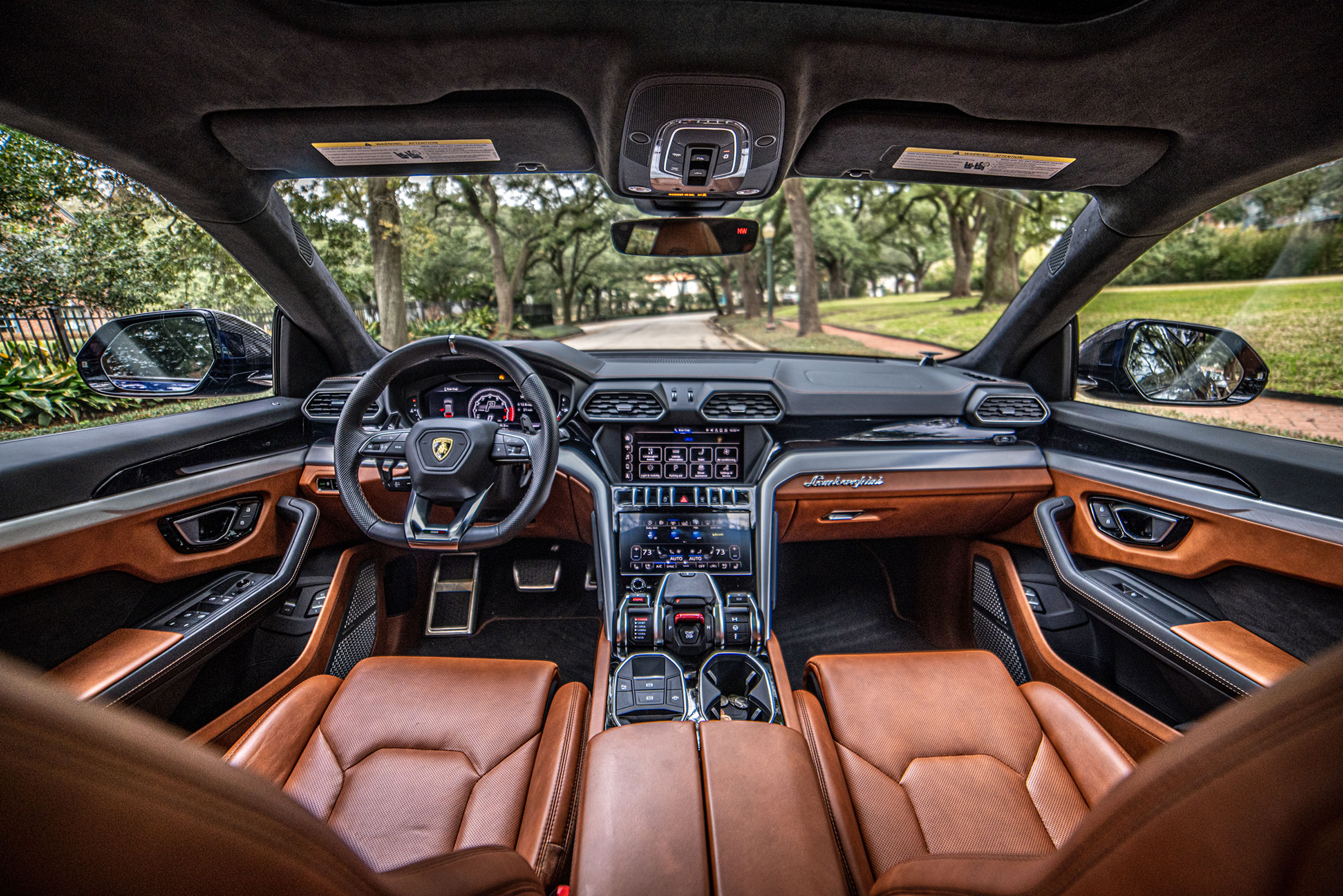 Interior view of a luxury car, a Lamborghini Urus, showcasing a sleek dashboard, high-tech controls, and brown leather seats with a scenic tree-lined street visible through the windshield.
